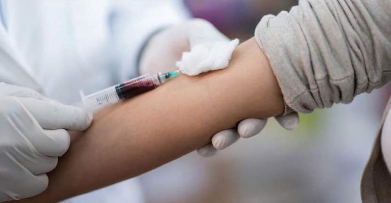 A gloved healthcare worker drawing blood from a patient's arm with a syringe and cotton swab at the injection site