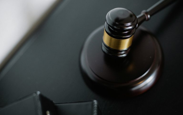 A wooden judge's gavel with a gold band resting on its sound block atop a dark surface