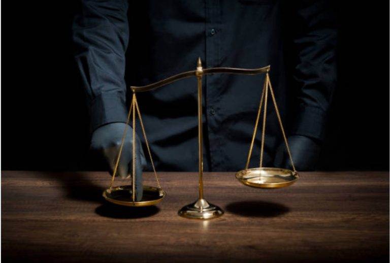 A gloved hand pressing down on one side of a gold balance scale, tipping it to suggest manipulation of justice