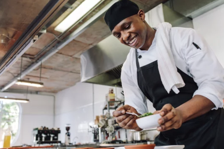 Smiling chef in a white coat and black apron plating a dish in a professional kitchen