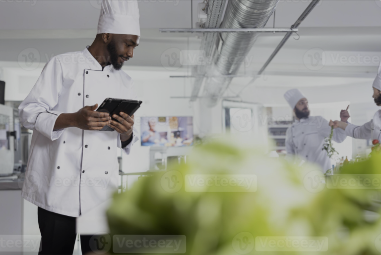 Chef in white uniform and toque reviewing a tablet in a professional kitchen, with other chefs working in the background
