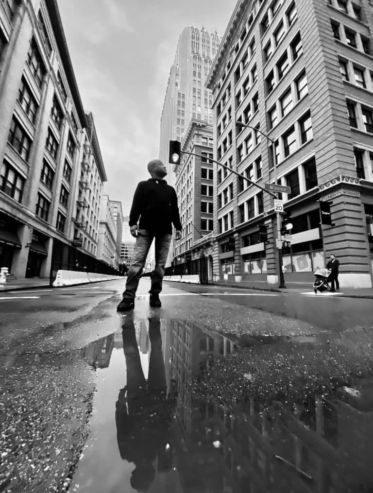Person standing at a downtown city intersection, reflected in a rain puddle, surrounded by tall buildings on an overcast day