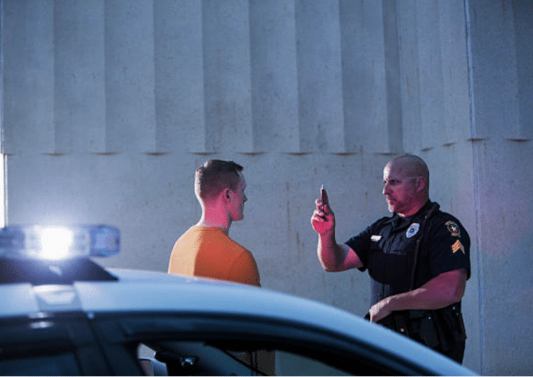 Police officer administering a field sobriety test to a man beside a patrol car with flashing lights
