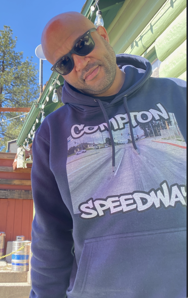 Bald man wearing sunglasses and a Compton Speedway graphic hoodie, standing outdoors on a sunny day